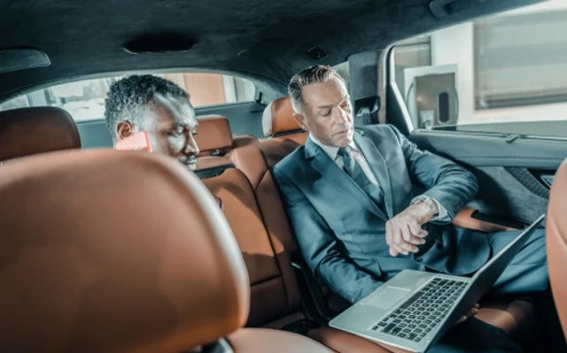 Two men in suits are seated in the back seat of a car, with one man engaged in a phone conversation and the other examining his wristwatch while holding an open laptop. This image depicts a scene of individuals being late, utilizing a black car service for luxury transportation, and relying on a dependable chauffeur.