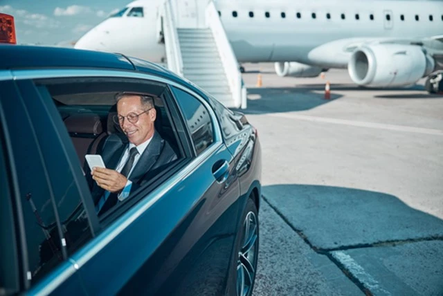Smiling businessman with glasses using cell phone during airport transfer after plane ride riding black car service.