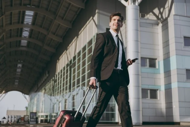 A young businessman in a black suit is walking outside the international airport terminal carrying a suitcase while talking on his mobile phone. He is booking a taxi and ordering a hotel for his business trip.