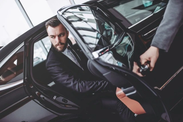 The young handsome businessman is sitting in a luxury car. A serious bearded man in a suit is coming out while the driver is opening the door using Corporate Transportation Services.