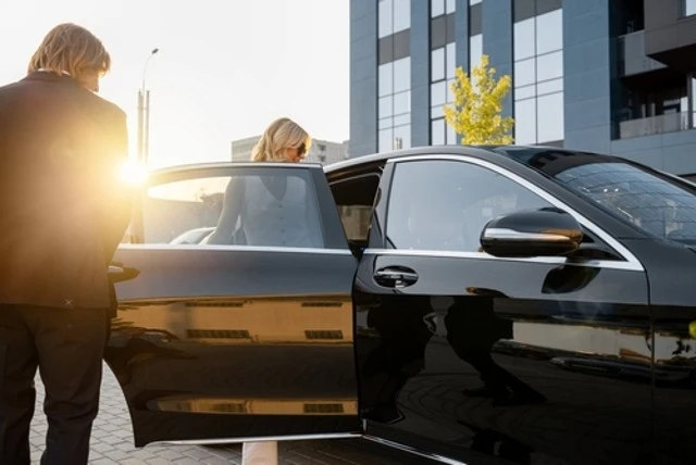 The chauffeur helps the business lady to get in the car, opening the vehicle door near the office building at sunset. Concept of business lifestyle and transportation, Your Business Travel Experience.