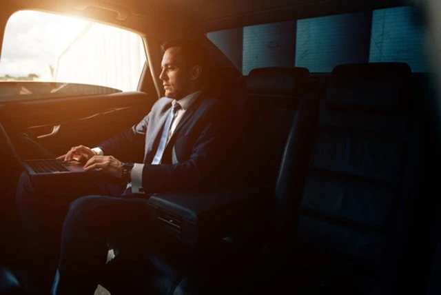 Handsome businessman talking with phone sitting with laptop in the backseat of the Black car service.