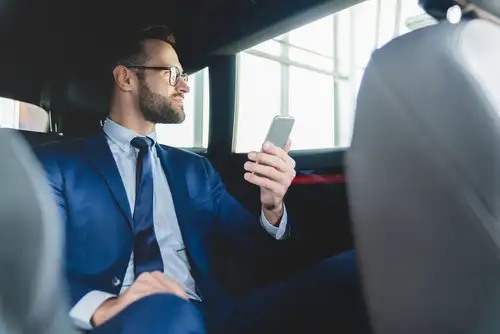 Experience luxury and convenience with our long distance ride service. Picture a confident businessman in a suit, using his smartphone in the backseat of a car.
