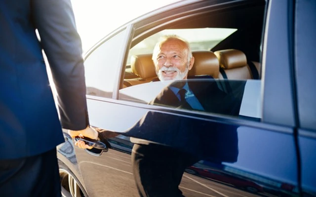 A professional chauffeur assists an elderly man in a suit as he exits a car, likely on his way to a medical appointment.
