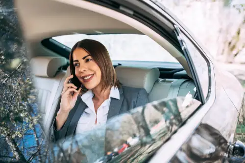 An elegant businesswoman in a car, engaged in a phone conversation. She sits comfortably in the backseat of a chauffeured limousine.